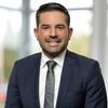 Ein Mann in einem blauen Anzug mit Krawatte lächelt in die Kamera. Der Hintergrund ist unscharf und zeigt ein Fenster.