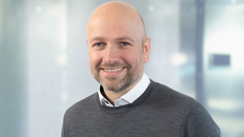Ein Porträt eines lächelnden Mannes in einem grauen Pullover vor einem unscharfen Hintergrund.