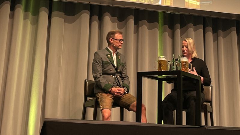 Auf der Bühne des Codex Netzwerk-Events 2025 sitzen zwei Personen in traditioneller bayerischer Kleidung und diskutieren. Im Hintergrund ist ein Plakat mit Münchner Wahrzeichen zu sehen.