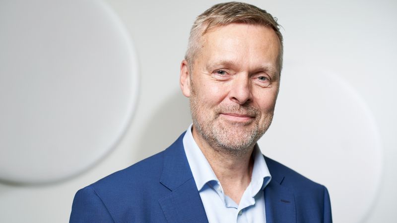 Ein Mann in einem blauen Anzug lächelt in die Kamera vor einem neutralen Hintergrund.