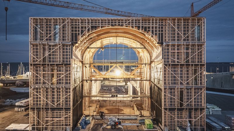Ein großes Holzgerüst auf einer Baustelle bei Nacht, beleuchtet von einem hellen Licht im Hintergrund.