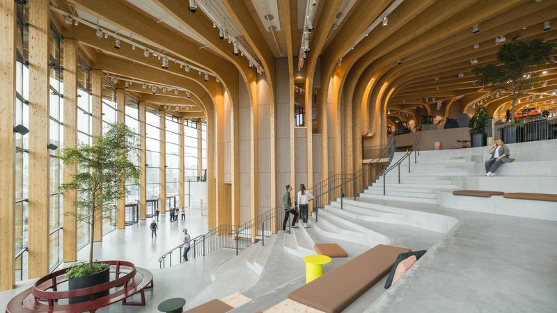 Ein moderner Innenraum eines Gebäudes mit beeindruckender Holzarchitektur und großen Fenstern, die viel Licht hereinlassen. Menschen bewegen sich durch den Raum, der mit Pflanzen und Sitzgelegenheiten ausgestattet ist.