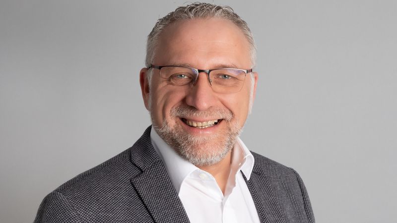 Ein Porträt eines lächelnden Mannes in einem grauen Sakko und weißem Hemd vor einem neutralen Hintergrund.