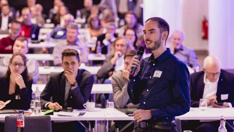 Ein Mann spricht in ein Mikrofon auf einer Konferenz vor einem Publikum.