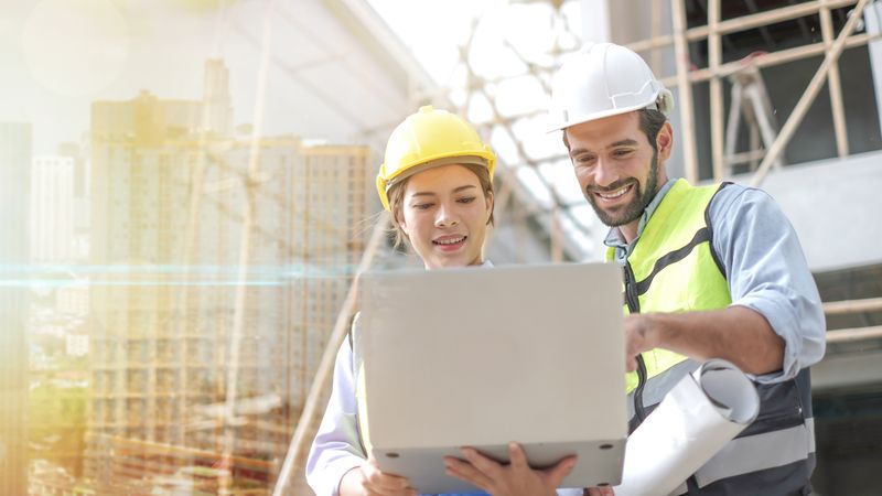 Zwei Bauarbeiter mit Schutzhelmen betrachten einen Laptop auf einer Baustelle.