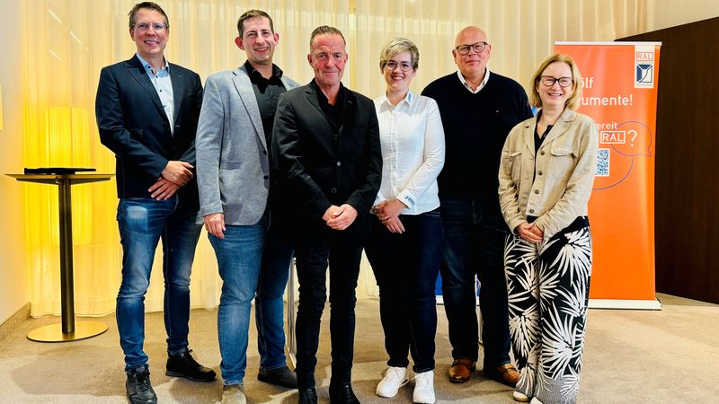 Eine Gruppe von sechs Personen steht vor einem orangefarbenen Banner in einem Konferenzraum.