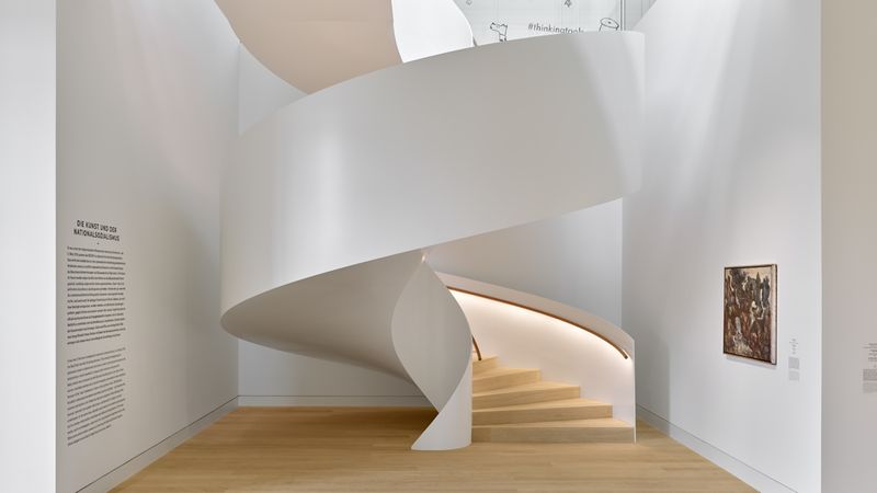 Eine moderne, weiße Wendeltreppe in einem Museum mit hölzernem Boden und Kunstwerken an den Wänden.