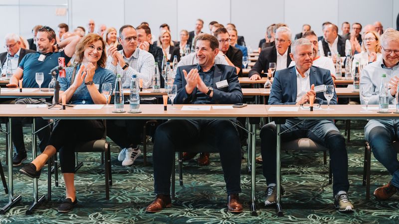 Teilnehmer einer Konferenz sitzen an Tischen und applaudieren.