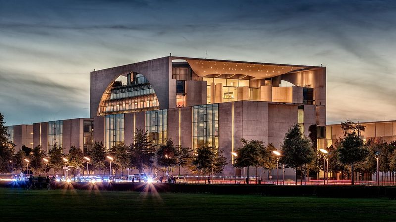 Das Bild zeigt das Bundeskanzleramt in Berlin während der Abenddämmerung, beleuchtet und umgeben von Bäumen.