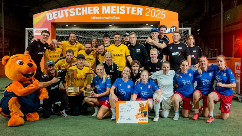 Eine Fußballmannschaft feiert den Titel "Deutscher Meister 2025" nach einem Elfmeterschießen. Die Spieler posieren mit einem Pokal und einem großen Scheck vor einem Tor.