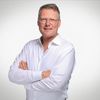 Ein Mann in einem weißen Hemd steht mit verschränkten Armen vor einem neutralen Hintergrund.