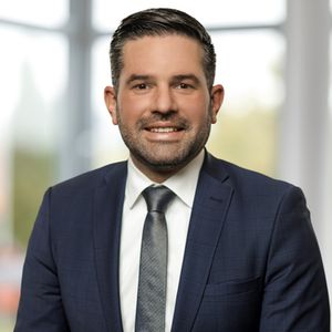 Ein Mann in einem blauen Anzug mit Krawatte lächelt in die Kamera. Der Hintergrund ist unscharf und zeigt ein Fenster.