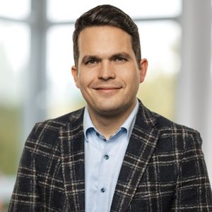 Ein Mann in einem karierten Sakko lächelt in die Kamera. Der Hintergrund ist unscharf und zeigt ein Fenster.