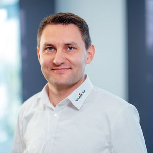 Ein Mann in einem weißen Hemd steht vor einem unscharfen Hintergrund und lächelt in die Kamera.