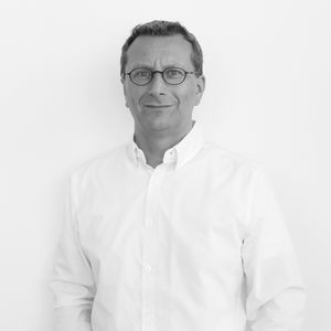 Ein Mann mit Brille in einem weißen Hemd steht vor einem neutralen Hintergrund.