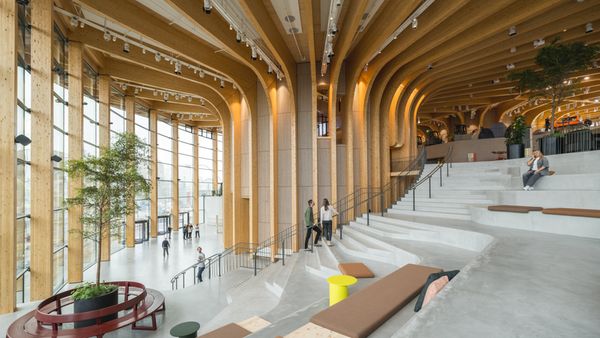 Ein moderner Innenraum eines Gebäudes mit beeindruckender Holzarchitektur und großen Fenstern, die viel Licht hereinlassen. Menschen bewegen sich durch den Raum, der mit Pflanzen und Sitzgelegenheiten ausgestattet ist.