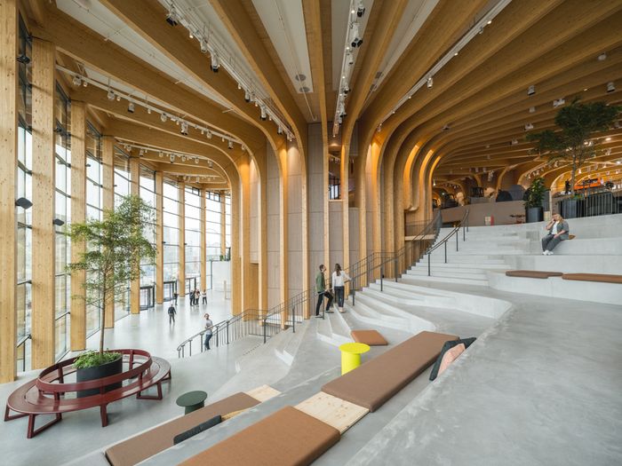 Ein moderner Innenraum eines Gebäudes mit beeindruckender Holzarchitektur und großen Fenstern, die viel Licht hereinlassen. Menschen bewegen sich durch den Raum, der mit Pflanzen und Sitzgelegenheiten ausgestattet ist.