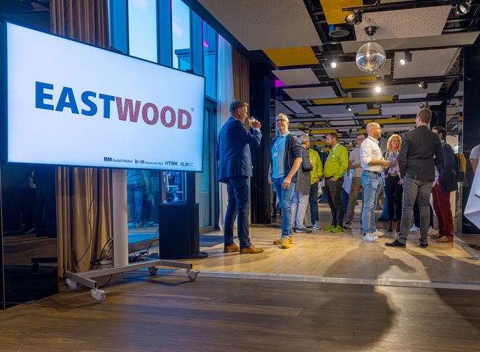 Ein Netzwerktreffen in einem modernen Büro mit mehreren Personen, die sich unterhalten und ein großes Display mit der Aufschrift "EASTWOOD".