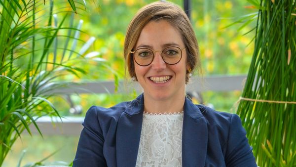 Eine lächelnde Frau mit Brille sitzt in einem Büro, umgeben von grünen Pflanzen.