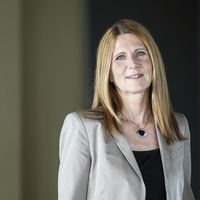 Eine professionelle Geschäftsfrau mit langen blonden Haaren, gekleidet in einem beige farbigen Blazer und einem schwarzen Oberteil, steht vor einem unscharfen Hintergrund, der ein Büro andeuten könnte.