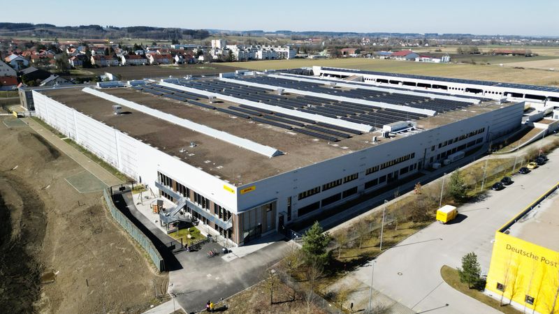 Luftaufnahme eines großen Logistikzentrums mit Solarpanelen auf dem Dach und umliegender Landschaft.