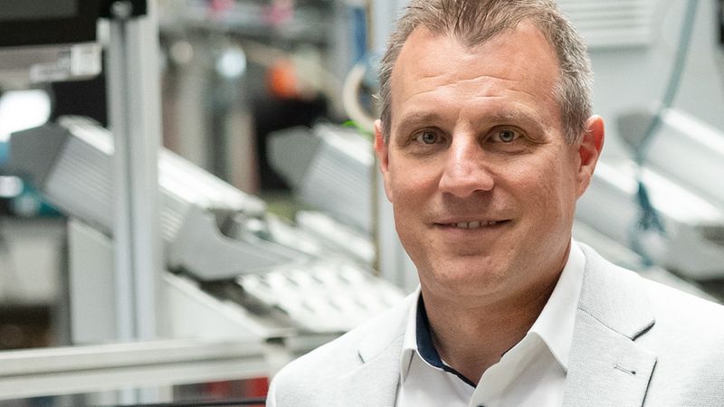 Ein Mann in einem grauen Anzug steht neben einem roten Rasenmäher in einer Fabrikumgebung.