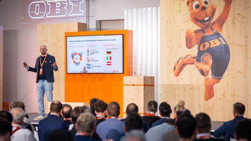 Ein Vortragender hält eine Präsentation auf einer Messe mit einem großen OBI-Logo und einem Maskottchen im Hintergrund.