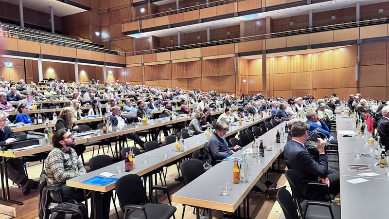 Ein großer Konferenzsaal voller Teilnehmer, die an einem Event teilnehmen. Die Teilnehmer sitzen an langen Tischen und hören aufmerksam zu.