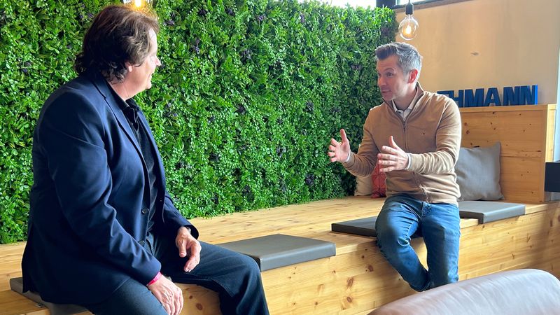 Zwei Männer führen ein Gespräch in einem modernen Büro mit einer grünen Pflanzenwand im Hintergrund.