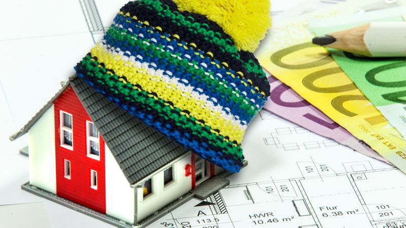 Ein Modellhaus mit einer bunten Wollmütze auf einem Stapel von Euro-Geldscheinen und Bauplänen.