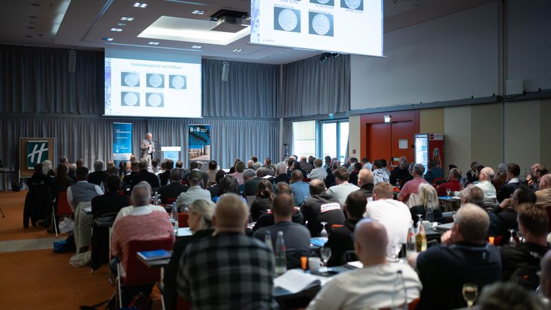 Ein Konferenzraum voller Teilnehmer, die einem Vortrag zuhören. Der Redner steht vorne und präsentiert Informationen auf einer Leinwand.