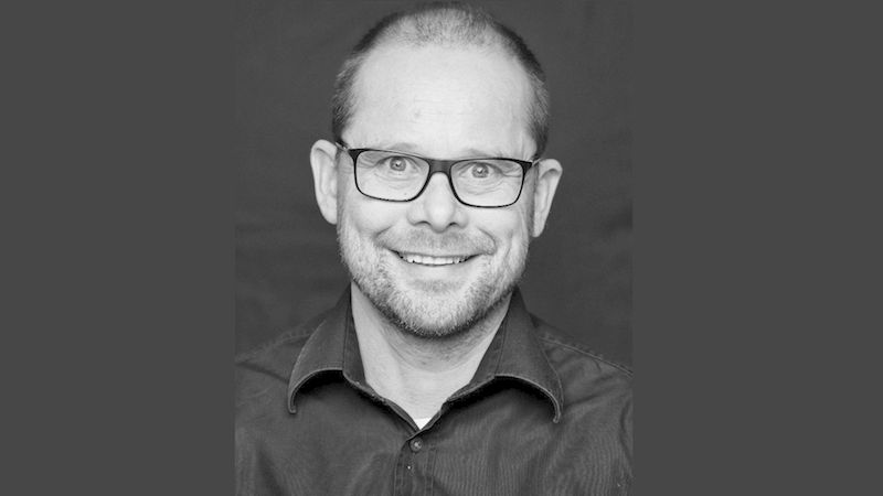 Dipl.-Ing. Kai Schulz. Ein schwarz-weiß Porträt eines lächelnden Mannes mit Brille vor einem dunklen Hintergrund.