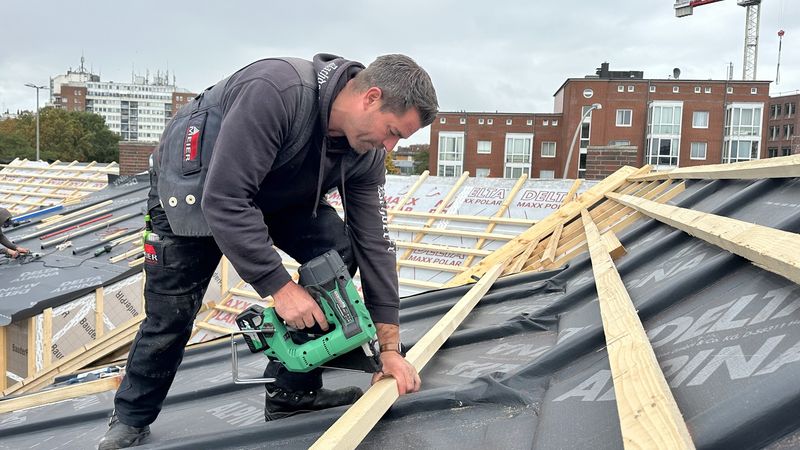 Ein Dachdecker arbeitet auf einem Dach und befestigt Holzlatten mit einem Nagelgerät.
