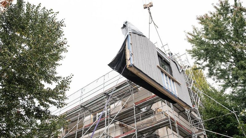 Ein Kran hebt ein Bauelement an einem Baugerüst empor, um es in ein Gebäude einzufügen.