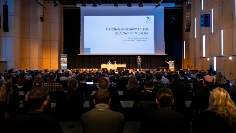 Eine große Konferenz in Münster mit vielen Teilnehmern, die einer Präsentation auf der Bühne folgen.