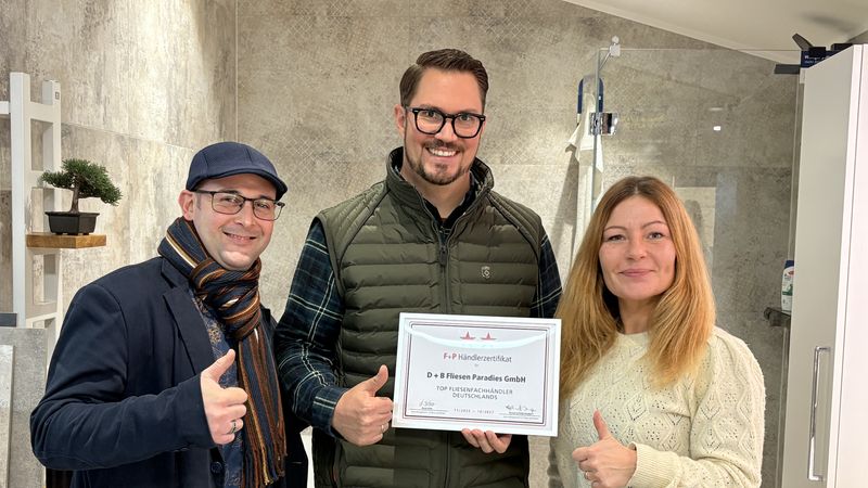 Drei Personen stehen in einem Raum und halten ein Zertifikat. Sie lächeln und zeigen Daumen hoch.