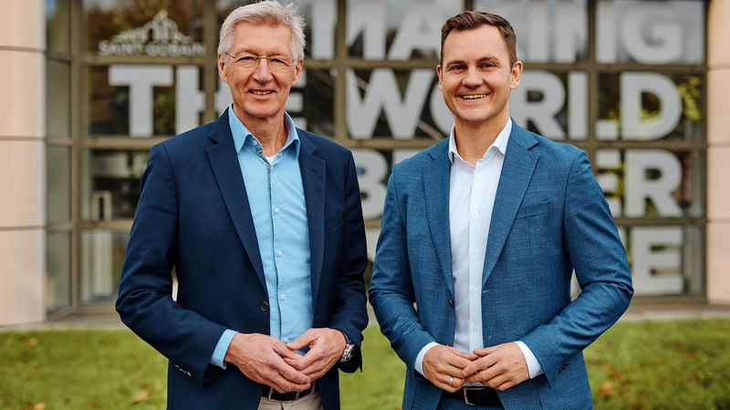 Zwei Männer in Anzügen stehen lächelnd vor einem modernen Gebäude mit Glasfassade.