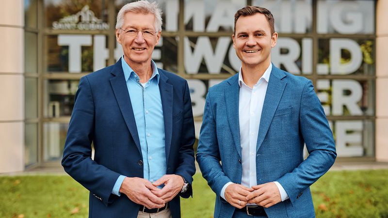 Zwei Männer in Anzügen stehen lächelnd vor einem modernen Gebäude mit Glasfassade.