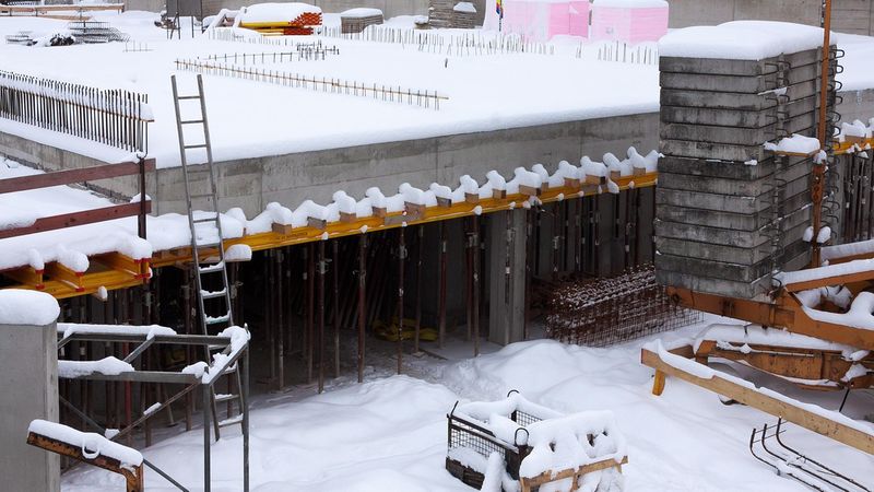 Eine Baustelle im Winter, bedeckt mit Schnee. Sichtbar sind Betonfundamente, Baugerüste und Baumaterialien, die unter einer Schneeschicht liegen.