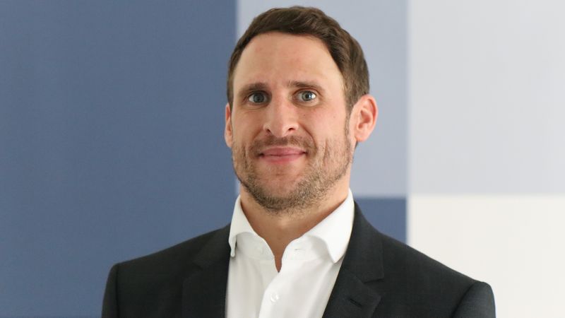 Ein Mann in einem Anzug vor einem blau-weißen Hintergrund.