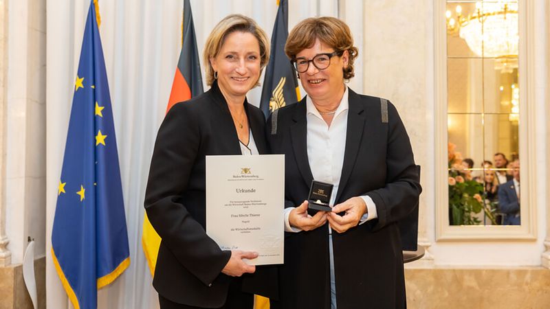 Zwei Frauen bei einer Auszeichnungszeremonie, eine hält eine Urkunde, die andere eine Medaille.