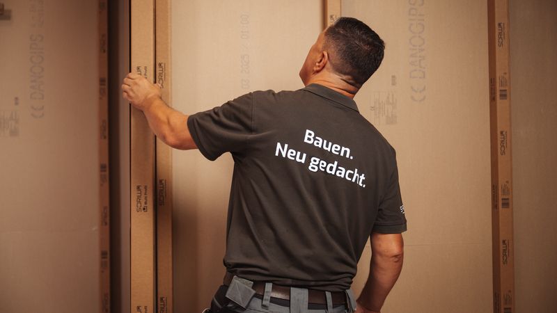 Ein Handwerker in einem dunklen T-Shirt steht vor einer Wand aus Gipskartonplatten und richtet diese aus.