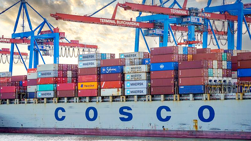 Ein großes Frachtschiff im Containerhafen, beladen mit zahlreichen Containern, während Kräne im Hintergrund zu sehen sind.