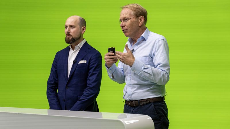 Zwei Männer stehen vor einem grünen Hintergrund. Einer hält ein Mikrofon und spricht, während der andere aufmerksam zuhört.