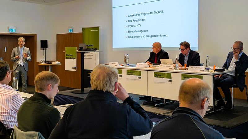 Ein Vortrag von Dr. Edgar Joussen vor den Teilnehmenden der Münchner Bauleitertage, bei dem die drei Mitreferenten auf einem Podium an einem Rednertisch sitzen.