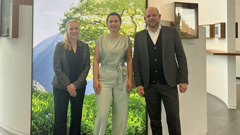Ein Gruppenfoto von drei Personen vor einem großen Display mit einem grünen Baum und dem Text "Wir denken Grün. Wir handeln Grün."