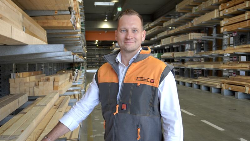 Ein Mitarbeiter steht in einem großen Holzlager und lächelt in die Kamera. Im Hintergrund sind Regale mit Holzplanken zu sehen.