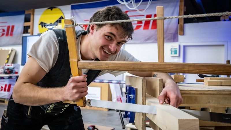 Ein Handwerker, der mit einer Säge an einem Holzstück arbeitet, lächelt in die Kamera.