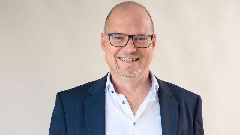 Ein lächelnder Mann in einem blauen Anzug mit weißem Hemd und Brille.