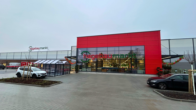 Der Eingang eines Hagebaumarkts mit auffälliger roter Fassade und geparkten Autos im Vordergrund.